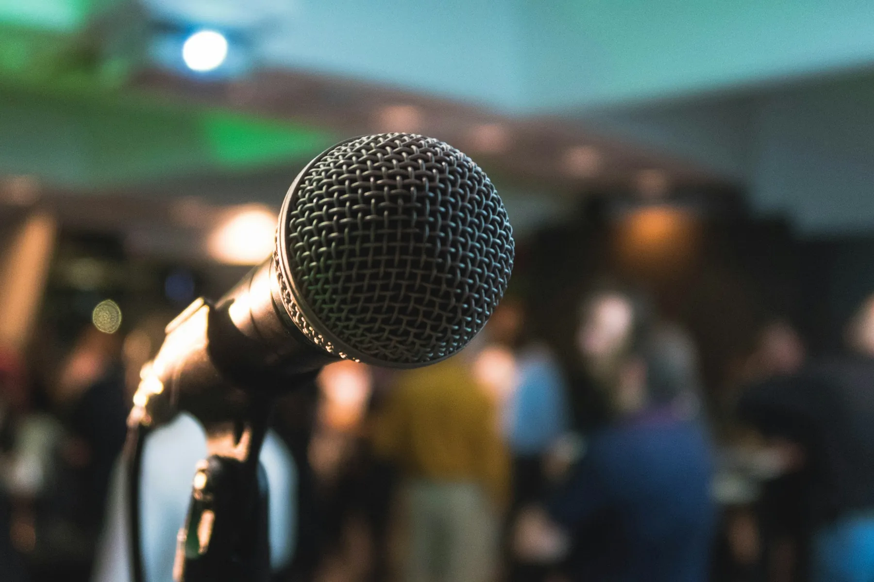 A microphone on a stand, lit from behind, with an out-of-focus audience filling the room beyond it