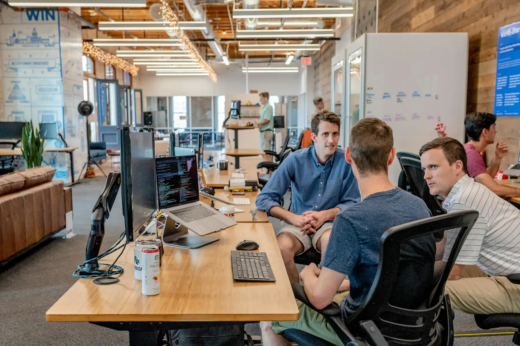 Three colleagues talking across a desk in an open-plan office, monitors and laptops around them